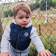 Tiago participe au concours pour gagner de l'argent avec cette photo : child, toddler, outdoor, fence, chain_link_fence, blue_eyes, casual_clothing, vest, hoodie, grass, motorbike, dirt_track, nature, person, portrait, curious, young_child, daylight, hands, background_blur