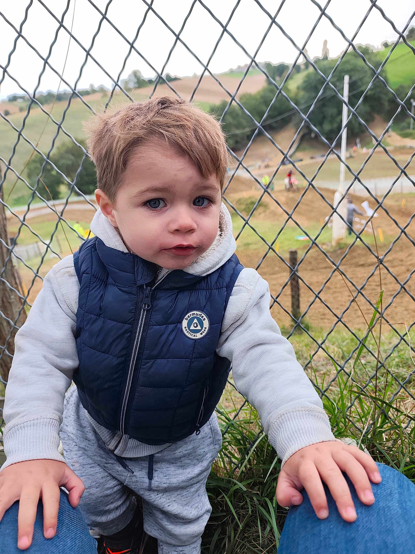 Tiago participe au concours pour gagner de l'argent avec cette photo : child, toddler, outdoor, fence, chain_link_fence, blue_eyes, casual_clothing, vest, hoodie, grass, motorbike, dirt_track, nature, person, portrait, curious, young_child, daylight, hands, background_blur