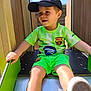 child, boy, slide, playground, outdoor, smiling, cap, sportswear, shorts, tshirt, happy, sunlight, toy, fun, recreation, daylight, person, feet, legs, green_clothing