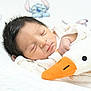 newborn, baby, sleeping, plush_toy, duck, soft_bedding, cartoon_background, child, infant, peaceful, cute, face, hand, white_clothing, cozy, indoors, portrait, resting, soft_texture, closeup
