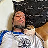 man, dog, sleeping, pillow, relaxation, indoor, casual_clothing, beard, pet, companion, resting, close_up, white_shirt, black_pillow, text_on_pillow, brown_dog, short_hair, necklace, comfortable, bond