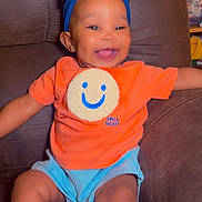 Love is registered to the contest to win money with this photo: baby, blue_headband, bright_colors, child, comfortable, couch, cute, expression, happy, headwear, indoor, infant, joyful, orange_shirt, person, portrait, shorts, sitting, smile, young_child