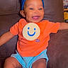 baby, child, smile, orange_shirt, blue_headband, couch, happy, indoor, person, sitting, cute, infant, bright_colors, headwear, shorts, portrait, young_child, comfortable, expression, joyful