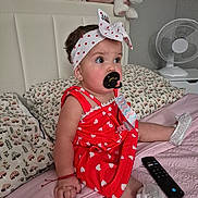 Daphne Nicolle is registered to the contest to win money with this photo: baby, child, red_dress, headband, pacifier, bed, pillow, plush_toys, remote_control, white_shoes, bracelet, indoor, curly_hair, cute, infant, sitting, person, soft_lighting, decor, room