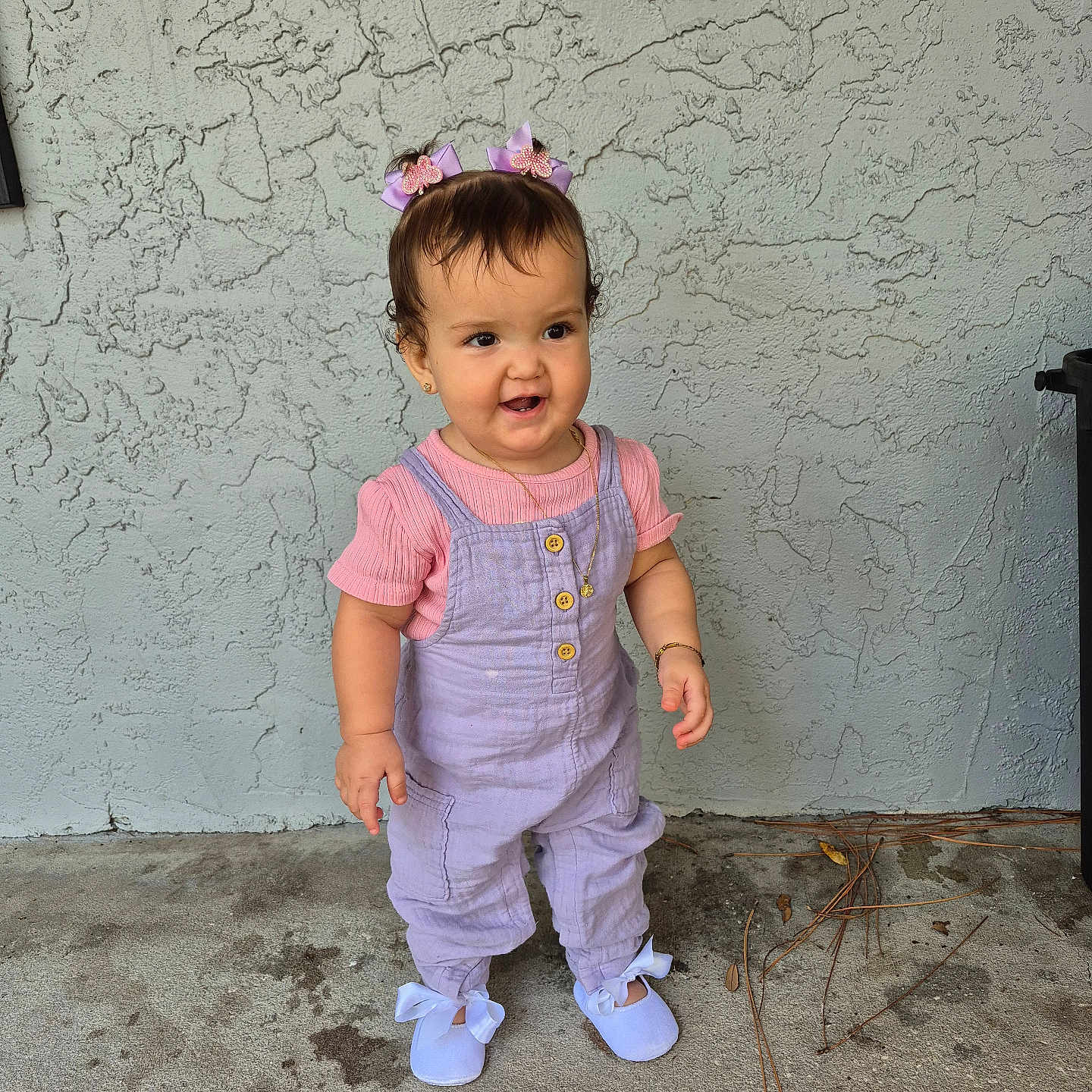 Daphne Nicolle is registered to the contest to win money with this photo: bracelet, casual_clothing, child, concrete_floor, curly_hair, cute, daylight, girl, hair_bows, necklace, outdoor, overalls, pink_shirt, ribbons, shoes, small_person, smiling, standing, textured_wall, toddler