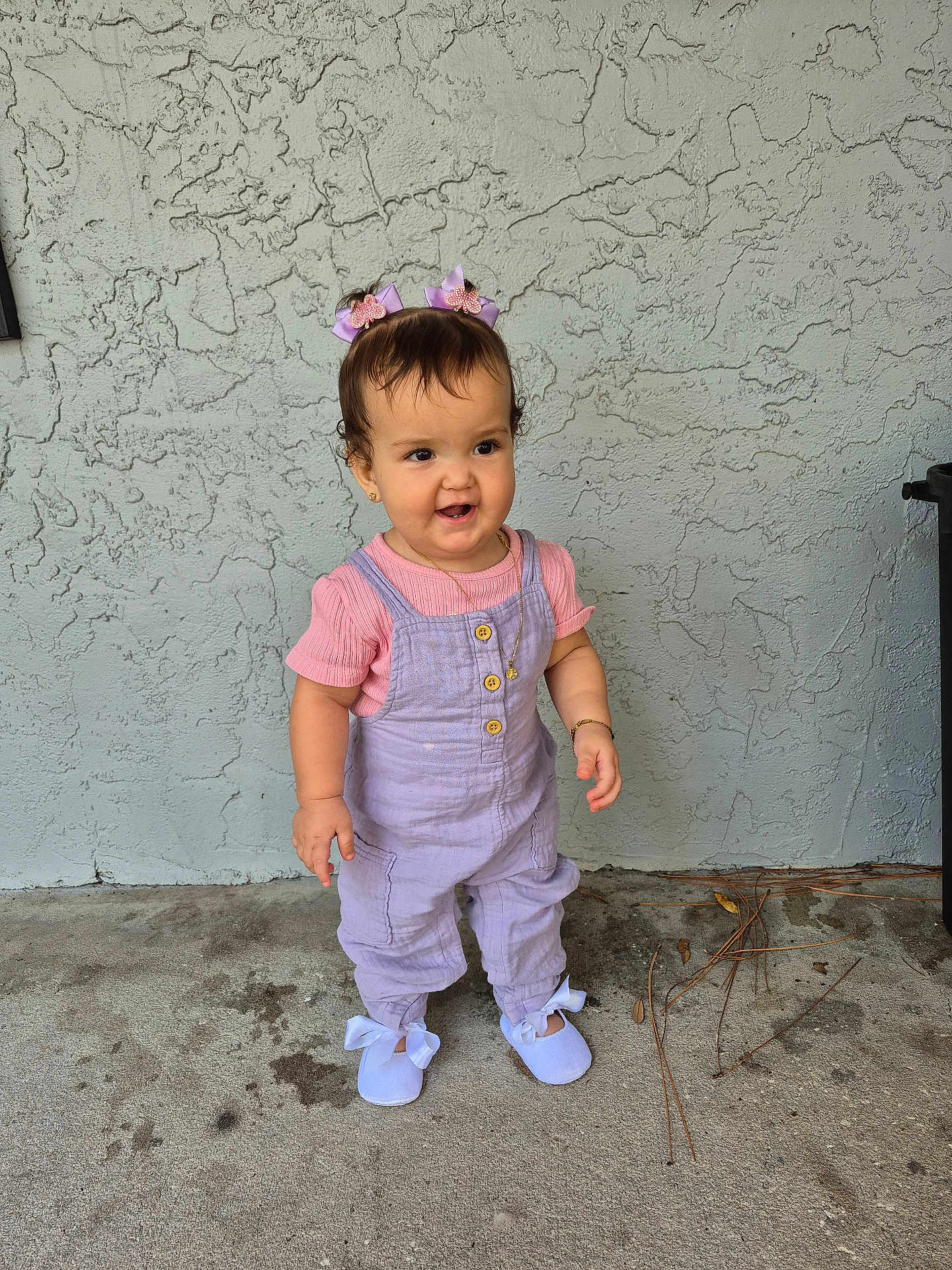 Daphne Nicolle is registered to the contest to win money with this photo: toddler, child, girl, standing, overalls, pink_shirt, hair_bows, shoes, ribbons, bracelet, necklace, concrete_floor, textured_wall, outdoor, curly_hair, cute, smiling, small_person, casual_clothing, daylight