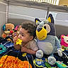 Amaris joined the competition — help win amazing prizes! baby, child, toy, plush, stuffed_animal, blanket, indoor, curly_hair, colorful, playpen, daylight, chewing, blue_dog, yellow_toy, furniture, window, vertical_blinds, person, infant, cute