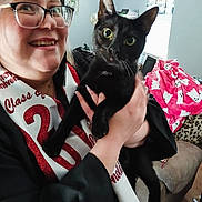 Sheba is registered to the contest to win money with this photo: person, cat, black_cat, graduation_gown, graduation_cap, sash, glasses, smile, indoor, living_room, blanket, window, furniture, hand, face, celebration, home, animal, pet, happy
