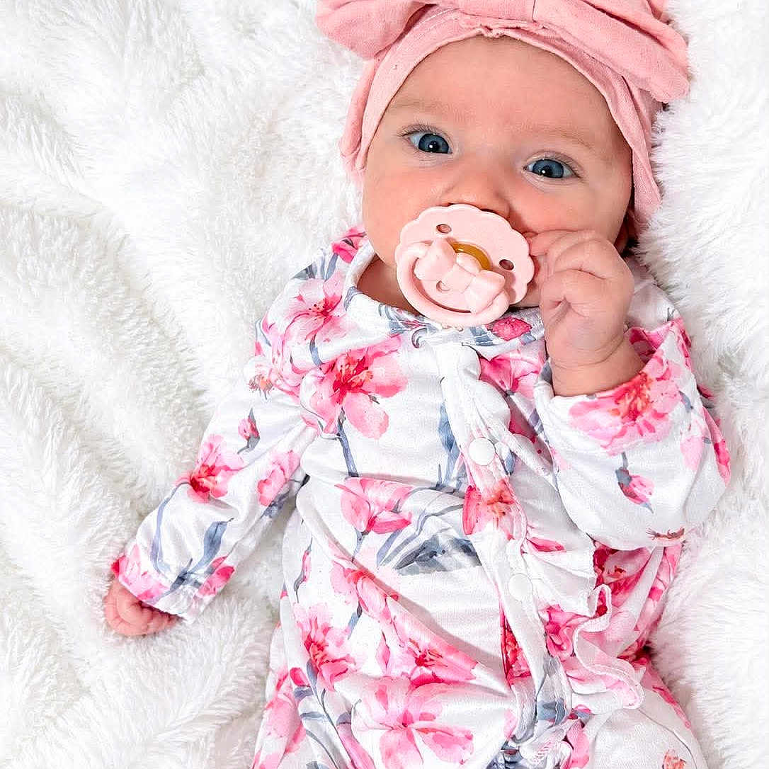Aria is registered to the contest to win money with this photo: baby, infant, child, portrait, floral_outfit, pink_clothing, headwrap, pacifier, smiling, blue_eyes, lying_down, blanket, white_background, soft_texture, indoor, closeup, fashion, adorable, hand, baby_photography