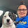 woman, dog, puppy, car, selfie, seatbelt, glasses, casual, indoor, portrait, smile, black_and_white_dog, blue_hoodie, short_hair, tie_dye_hair_tie, seat, window, daylight, closeup, companion