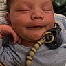 Dawson is registered to the contest to win money with this photo: baby, pacifier, infant, sleeping, smile, clothing, blue_outfit, hand, face, head, indoor, person, closeup, cute, resting, newborn, peaceful, soft_light, beads, background