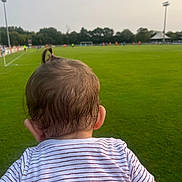 Ysalia participe au concours pour gagner de l'argent avec cette photo : toddler, child, back_of_head, hair, ponytail, striped_shirt, grass, soccer_field, sports, outdoor, daytime, people, green, blurred_background, stadium, trees, goalpost, playing_field, spectator, casual_clothing
