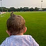 toddler, child, back_of_head, hair, ponytail, striped_shirt, grass, soccer_field, sports, outdoor, daytime, people, green, blurred_background, stadium, trees, goalpost, playing_field, spectator, casual_clothing