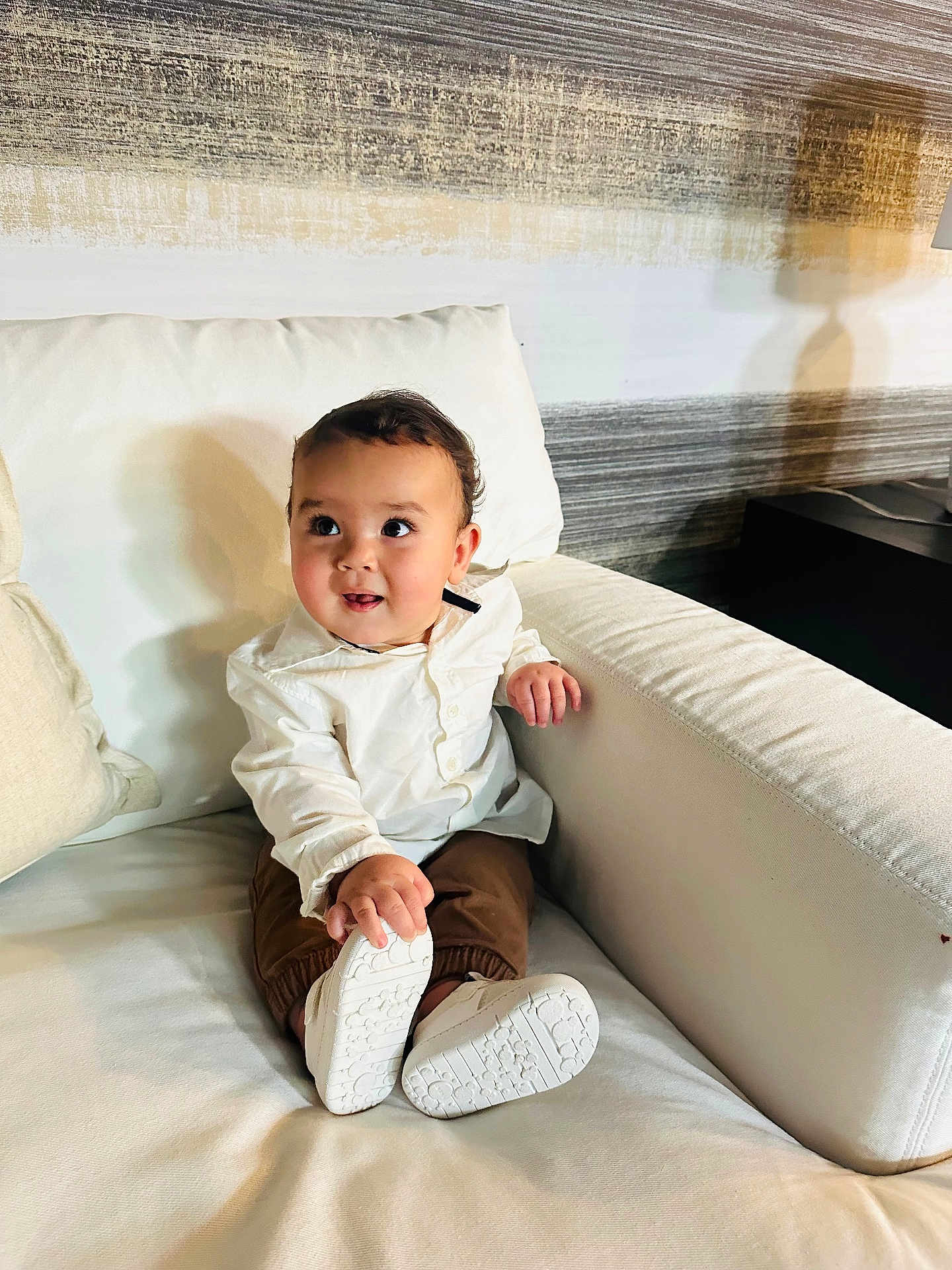 Mathius Durán joined the competition — help win amazing prizes! baby, toddler, child, white_shirt, brown_pants, white_shoes, couch, living_room, indoor, curious_expression, sitting, furniture, home, cute, person, portrait, young_child, smiling, comfortable, cozy