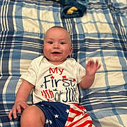 Mikey is registered to the contest to win money with this photo: baby, bedspread, blue, celebration, child, clothing, face, hand, happy, indoor, lying_down, patriotic, pattern, person, plaid, red, shirt, shorts, smiling, white