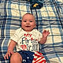 baby, bedspread, blue, celebration, child, clothing, face, hand, happy, indoor, lying_down, patriotic, pattern, person, plaid, red, shirt, shorts, smiling, white