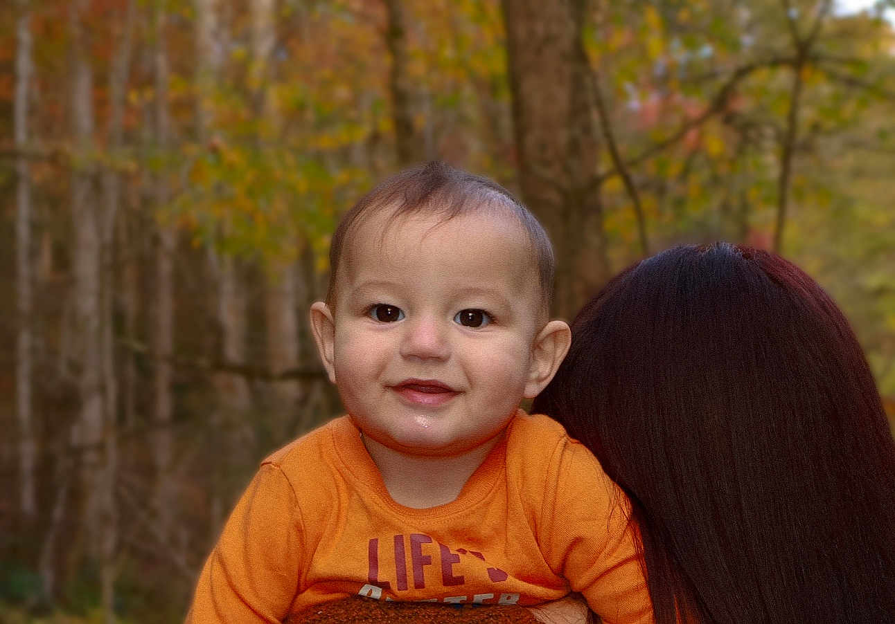 Liam is registered to the contest to win money with this photo: baby, child, smiling, orange_shirt, person, forest, trees, autumn, outdoor, nature, portrait, head, face, happy, young, holding, hair, background, season, cute
