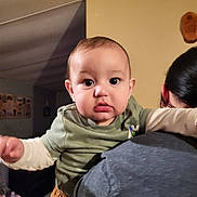 Liam is registered to the contest to win money with this photo: baby, child, person, indoor, holding, shoulder, curious, wide_eyes, chubby_cheeks, long_sleeves, green_shirt, brown_pants, adult, hair, face, portrait, home, wall, lighting, expression