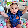 toddler, child, ball_pit, colorful_balls, blue_shirt, embroidery, playful_expression, indoor, bookshelf, children_books, baby_supplies, diapers, toy, happy, reaching_out, playroom, cute, portrait, person, smiling