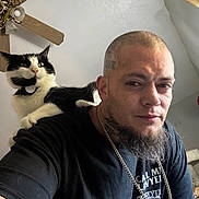 Pippy joined the competition — help win amazing prizes! person, cat, black_and_white_cat, pet, selfie, portrait, beard, tattoo, necklace, tshirt, indoor, ceiling_fan, living_room, shoulder_perch, fur, collar, eyes, wall, expression, head