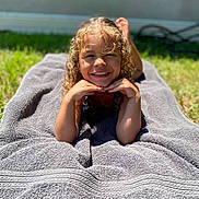 Ava is registered to the contest to win money with this photo: child, smiling, curly_hair, towel, outdoor, sunlight, grass, happy, portrait, young, relaxing, person, casual, daylight, skin, head, face, barefoot, nature, leisure