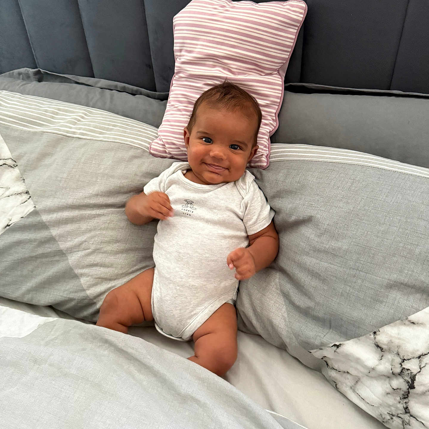 Shyro is registered to the contest to win money with this photo: baby, bed, child, cozy, cute, gray_bedding, happy, headboard, indoors, infant, marble_pattern, onesie, person, pillow, portrait, relaxed, resting, smile, striped_pillow, young