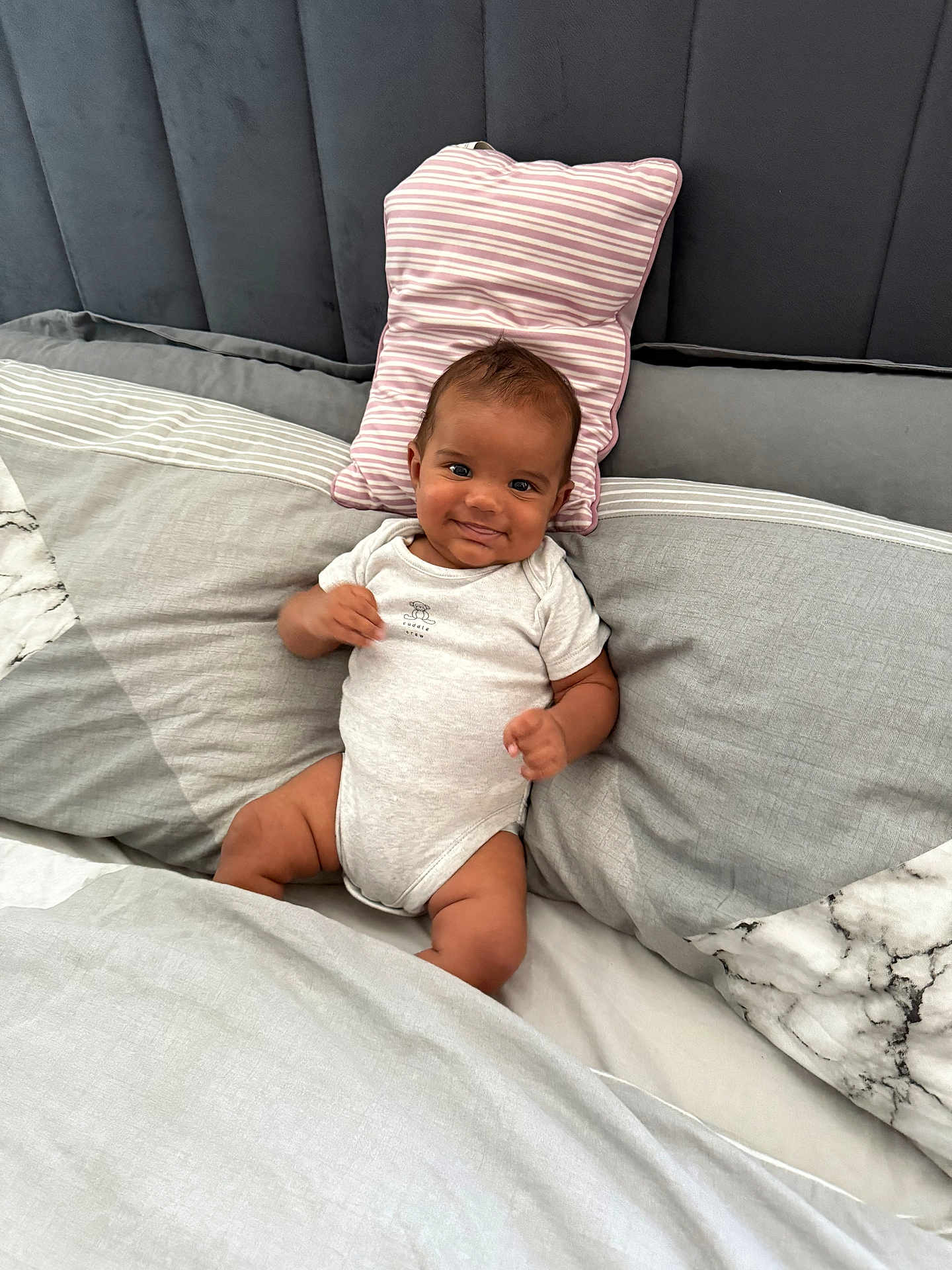 Shyro is registered to the contest to win money with this photo: baby, infant, smile, onesie, bed, pillow, striped_pillow, gray_bedding, marble_pattern, headboard, cute, child, happy, indoors, portrait, relaxed, young, person, resting, cozy