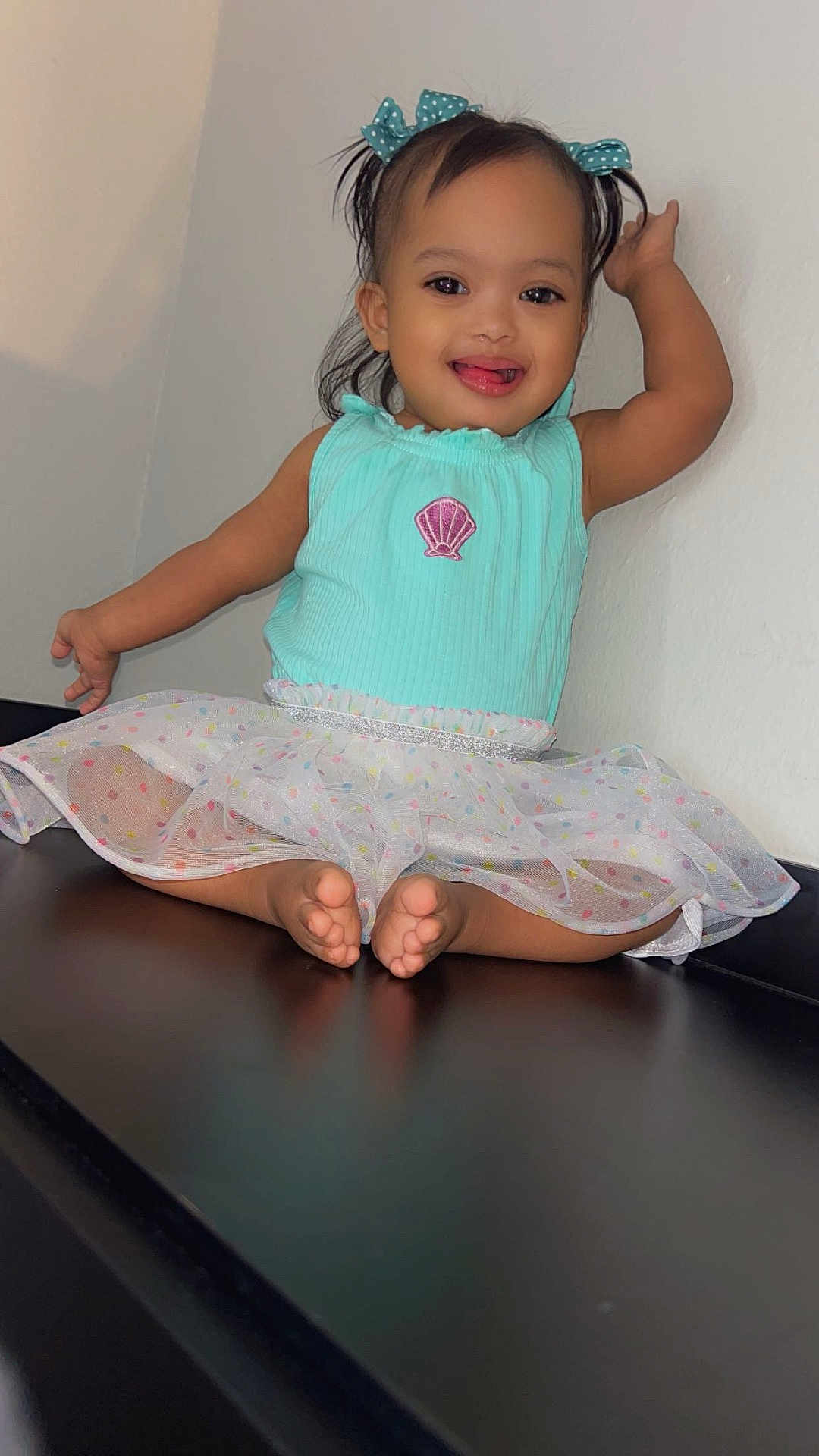 Emely is registered to the contest to win money with this photo: toddler, child, smiling, pigtails, hair_bows, mint_green_top, seashell_emblem, polka_dot_tutu, barefoot, sitting, dark_surface, cheerful, playful, indoor, wall, cute, happy, young_child, portrait, innocence