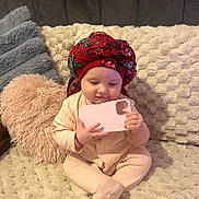Zenayda is registered to the contest to win money with this photo: baby, infant, pajamas, headscarf, red, pink, phone_case, blanket, couch, pillow, fluffy, soft, texture, cute, child, sitting, indoors, cozy, person, portrait