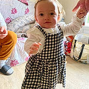 Solyna participe au concours pour gagner de l'argent avec cette photo : toddler, child, indoor, walking, checkered_dress, holding_hand, living_room, toy, floor, curly_hair, person, couch, blanket, socks, adult_hand, window, bag, furniture, flooring, curiosity