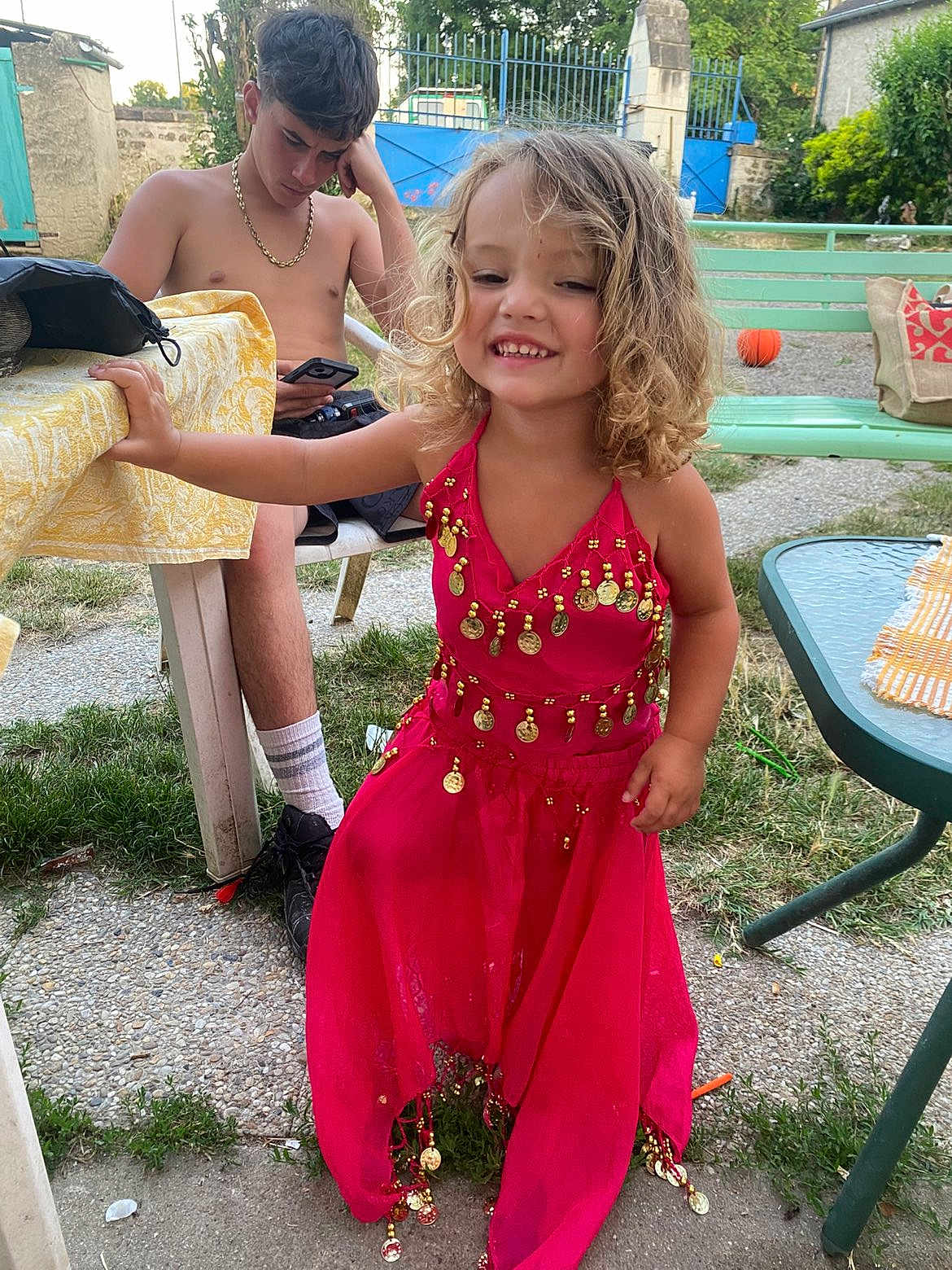 Solyna participe au concours pour gagner de l'argent avec cette photo : child, girl, smiling, dress, pink_dress, gold_coins, curly_hair, outdoor, grass, bench, table, young_man, shirtless, phone, chain, happy, summer, casual, person, playful