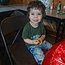 child, toddler, smiling, curly_hair, green_shirt, blue_jeans, folding_chair, indoor, table, birthday_gift_bag, soda_cans, toys, party, happy, brown_chair, wooden_wall, red_balloon, person, casual_clothing, celebration