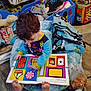 toddler, child, book, reading, colorful, toy, blanket, cushion, floor, indoor, learning, curly_hair, feet, play_area, plastic_bag, wooden_floor, clothing, early_education, person, cozy