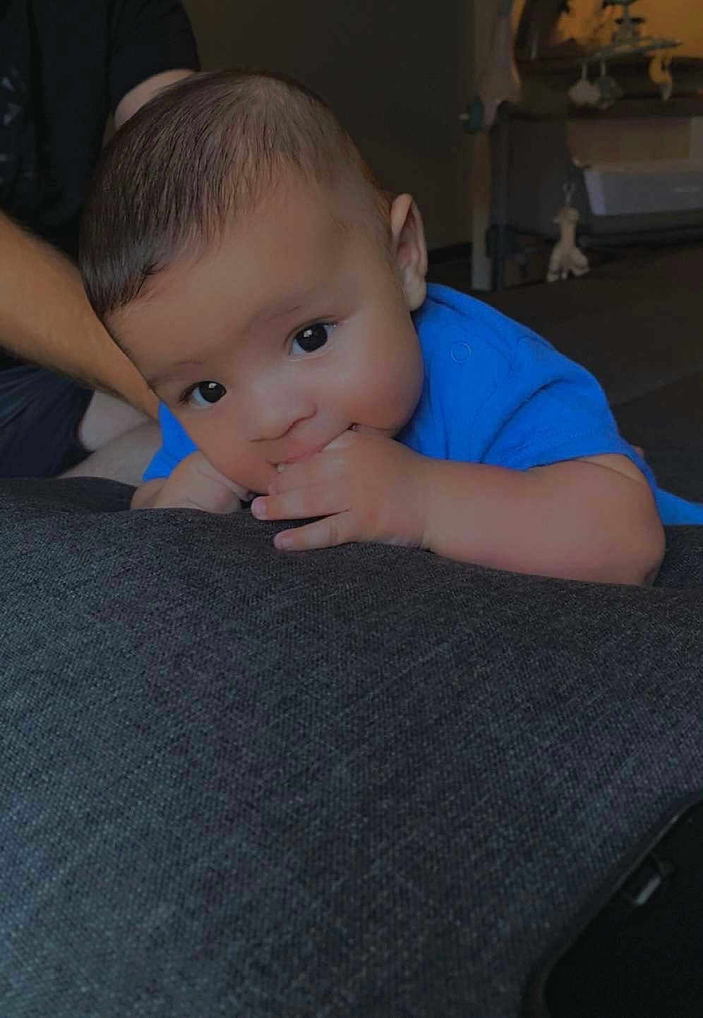 Zayan participe au concours pour gagner de l'argent avec cette photo : baby, infant, blue_shirt, couch, fingers, hand, face, person, indoors, curious, young_child, skin, hair, adult_arm, support, lying_down, close_up, expression, comfort, soft_lighting