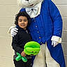 balloon, black_jacket, blue_coat, bow_tie, bunny, celebration, child, costume, curly_hair, event, friendship, green_balloon, indoor, mascot, person, portrait, smile, toy, wall, white_gloves