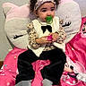 black_pants, cat_pillow, child, colorful, curly_hair, cute, expression, headband, indoor, minnie_mouse, pacifier, pink_blanket, plush_pillow, portrait, seated, socks, soft_toys, toddler, wall, white_vest