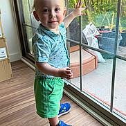 Makai is registered to the contest to win money with this photo: blue_shirt, blue_sneakers, casual_clothing, child, cute, daylight, glass_door, green_shorts, hand_raised, happy, indoor, person, portrait, smiling, standing, sunlight, toddler, window, wooden_floor, young_child