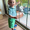 blue_shirt, blue_sneakers, casual_clothing, child, cute, daylight, glass_door, green_shorts, hand_raised, happy, indoor, person, portrait, smiling, standing, sunlight, toddler, window, wooden_floor, young_child