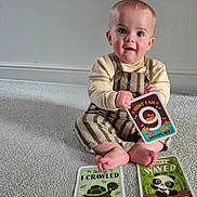 Kaladin is registered to the contest to win money with this photo: baby, child, infant, milestone_card, carpet, sitting, smiling, cute, clothing, striped_overalls, milestone, indoor, floor, person, young_child, happy, face, hands, feet, toy_card