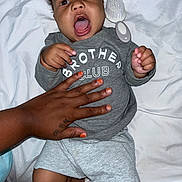 Blessingmoses is registered to the contest to win money with this photo: baby, child, infant, person, hand, tattoo, clothing, shirt, grey, shorts, white_sheet, brush, nail_polish, orange_nails, expression, open_mouth, lying_down, skin, indoor, cozy