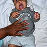 baby, child, infant, person, hand, tattoo, clothing, shirt, grey, shorts, white_sheet, brush, nail_polish, orange_nails, expression, open_mouth, lying_down, skin, indoor, cozy