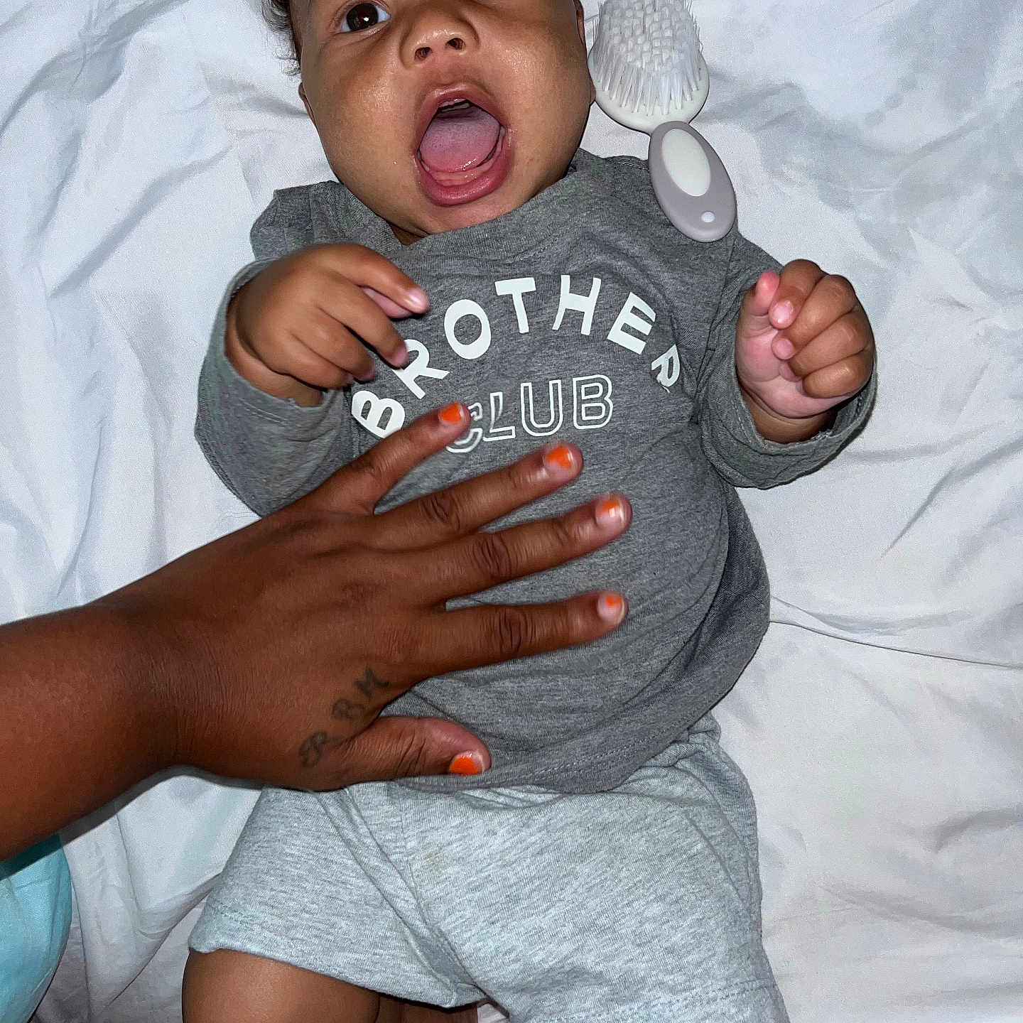 Blessingmoses is registered to the contest to win money with this photo: baby, child, infant, person, hand, tattoo, clothing, shirt, grey, shorts, white_sheet, brush, nail_polish, orange_nails, expression, open_mouth, lying_down, skin, indoor, cozy