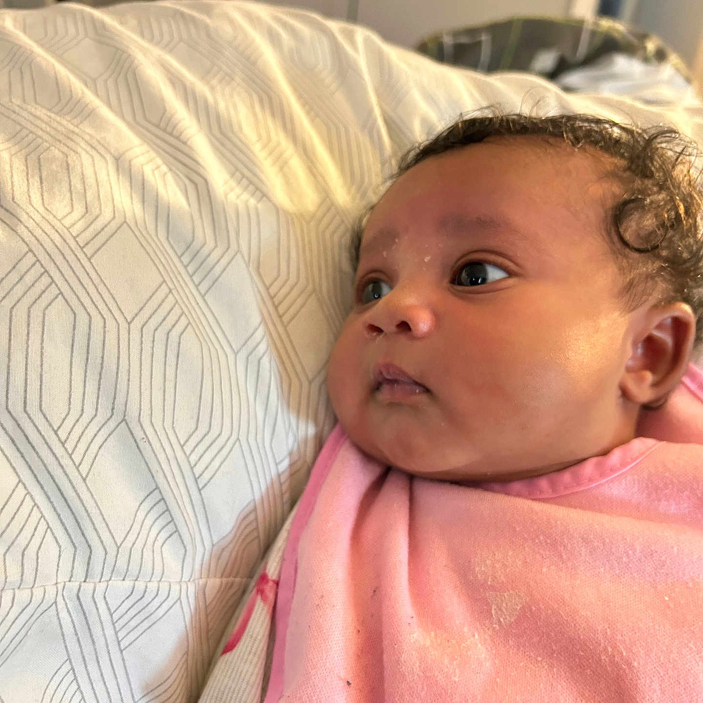 Nariah is registered to the contest to win money with this photo: baby, infant, face, curly_hair, pink_blanket, bed, bedding, sheet, pattern, indoor, child, person, skin, portrait, cute, newborn, lying_down, closeup, soft_light, expression