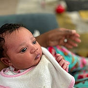 Nariah is registered to the contest to win money with this photo: baby, infant, close_up, bib, pink_trim, curly_hair, hand, adult_hand, indoor, soft_focus, colorful_background, person, cute, newborn, portrait, tender_moment, skin, clothing, sleepwear, expression