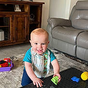 Rayden joined the competition — help win amazing prizes! toddler, child, smiling, indoor, living_room, toy, television, couch, carpet, wooden_furniture, frame, happy, play, person, floor, green_toy, blue_clothing, bib, plastic_toy, home