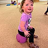 black_pants, brown_boots, carpeted_floor, casual_clothing, child, colorful_mats, curly_hair, indoor, other_children, panda_shirt, person, play_area, playful, purple_ball, rosy_cheeks, sitting, smiling, support_pole, toddler, wooden_door