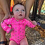 baby, child, pink_swimsuit, polka_dots, outdoor, sunlight, curious_expression, blue_eyes, holding_hands, legs, feet, chain_link_fence, leaves, concrete_ground, adult_hands, sandals, skin, summer, playtime, casual