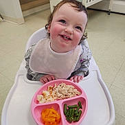 Navaya is registered to the contest to win money with this photo: toddler, child, high_chair, bib, food, plate, shredded_chicken, green_beans, mandarin_oranges, smiling, indoor, crib, stairs, floor, happy, eating, curly_hair, person, meal, pink_plate
