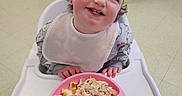 Navaya is registered to the contest to win money with this photo: bib, child, crib, curly_hair, eating, floor, food, green_beans, happy, high_chair, indoor, mandarin_oranges, meal, person, pink_plate, plate, shredded_chicken, smiling, stairs, toddler
