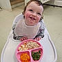 toddler, child, high_chair, bib, food, plate, shredded_chicken, green_beans, mandarin_oranges, smiling, indoor, crib, stairs, floor, happy, eating, curly_hair, person, meal, pink_plate